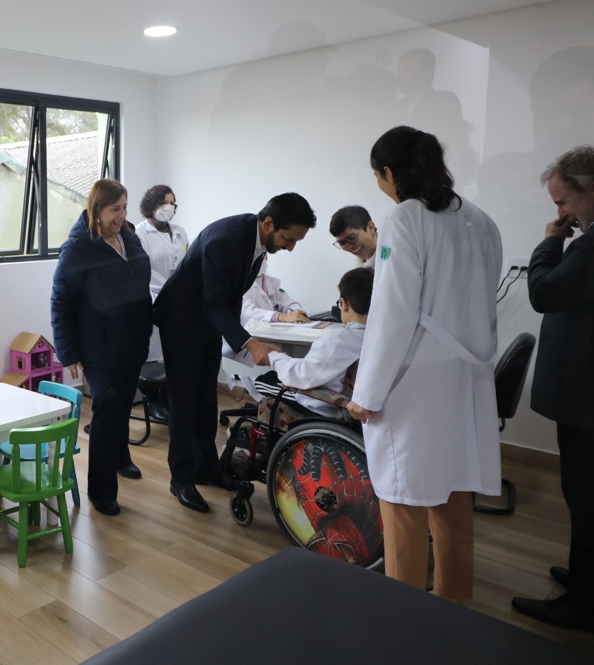 Reunidos em uma das salas de atendimento do CER, o prefeito Ricardo Nunes com a secretária municipal da SMPED, Silvia Grecco. Eles conversam com uma mãe e o filho com deficiência, ele usa uma cadeira de rodas.